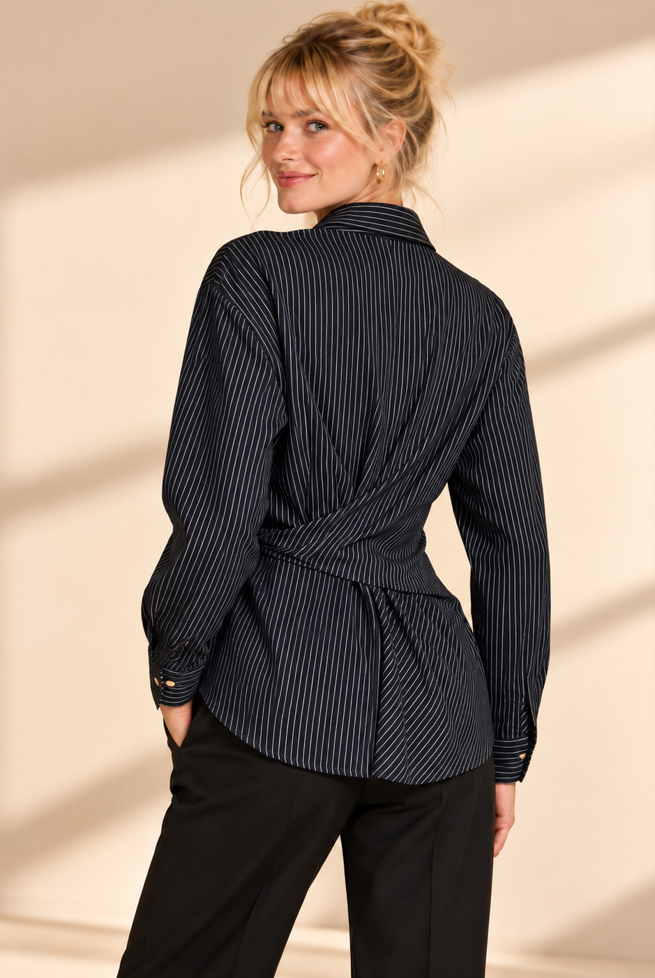 Svart blus Ester med smala ljusa ränder, skjortkrage, guldfärgade knappar och knytband i midjan samt långa ärmar med manschett, visad på modell.